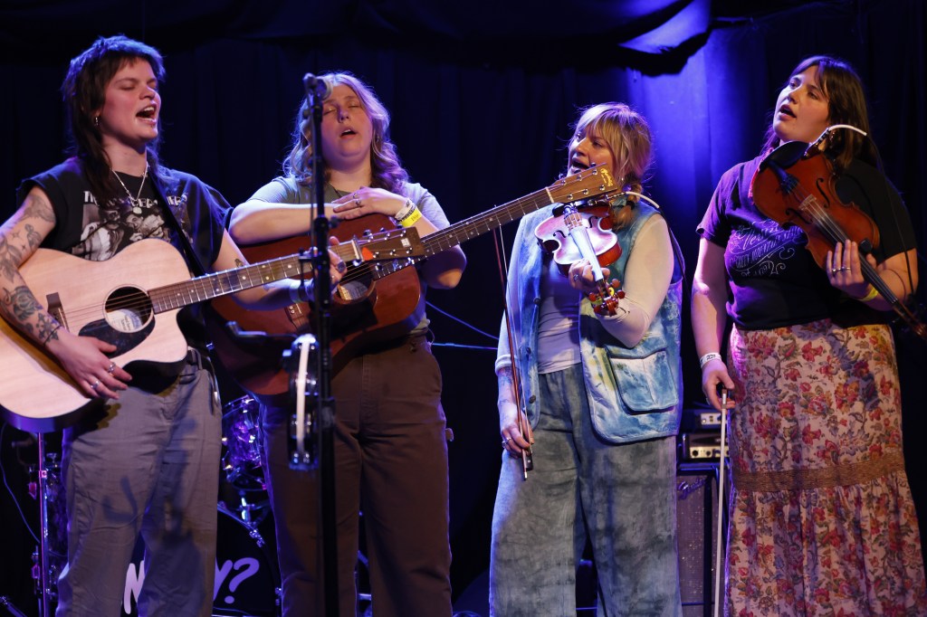 Four of Bitch Creek's five members are shown gathered around a microphone, singing in harmony. To the right of the image are two fiddle players, and to the left are two acoustic guitarists. Only their upright bass player is out of frame. 