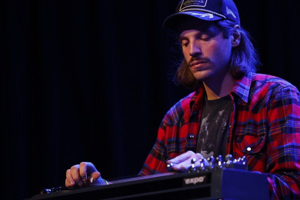 The Spine Stealers' James Strelow is pictured in a baseball cap and red flanel, playing his pedal steel guitar, an instrument that runs along the bottom frame of the image at a diagonal. His eyes are closed, and he is sitting down, appearing mostly to the far right of the image, though his fingerpicking hand is in the bottom left corner.  