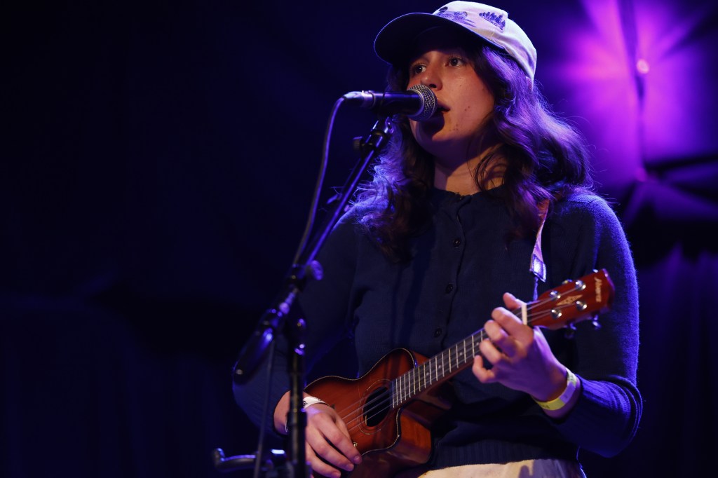 The Spine Stealers' Emma O'Shea plays ukulele while singing into the microphone. She's wearing a yellow wristband, baseball cap, and a dark long-sleeved shirt.