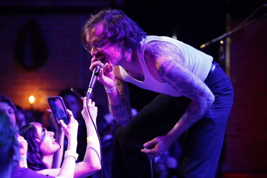Excuse Me, Who Are You? vocalist Kyle Kinney performs at the front of the stage, bent over and screaming into the mic directly in front of the crowd. A woman in the bottom right is seen smiling with her phone held up, taking video of the moment. Another phone is also visibly recording in the same section. 